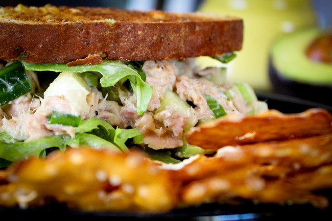 Toasted whole wheat toast, with tuna salad, avocado sliced and arugula. Avocado half in the background and thin pretzels in the foreground. 