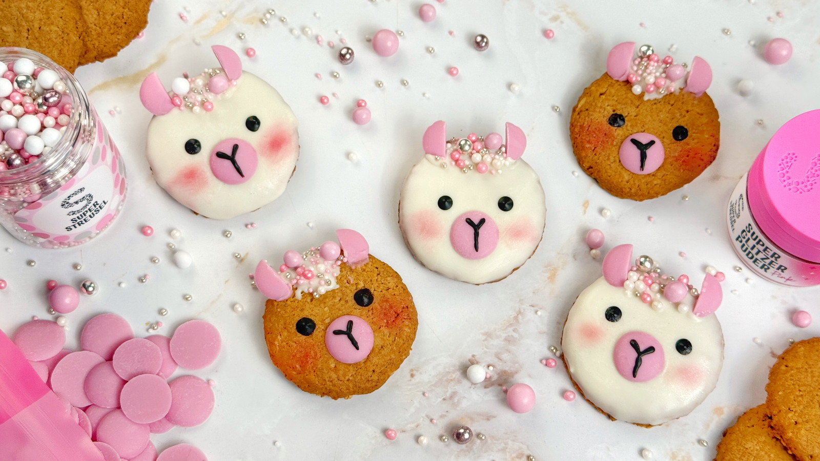 Alpaka-Kekse aus Haferkeksen, dekoriert als weiße und braune Alpaka-Gesichter mit weißem und schwarzem SuperDrip, rosa SuperDrops, StreuselMix KuschelMuschel als Wolle und pinkem SuperGlitzerPuder auf den Bäckchen.