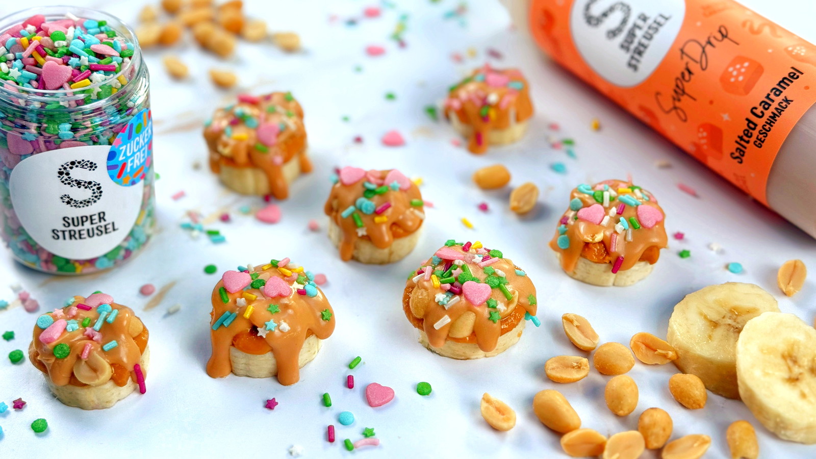 Banaan-pinda bites van verse bananenschijfjes met pindakaas en gezouten pinda’s, afgewerkt met Salted Caramel SuperDrip en suikervrije StreuselMix PartyExplosion. Snelle snack zonder bakken.