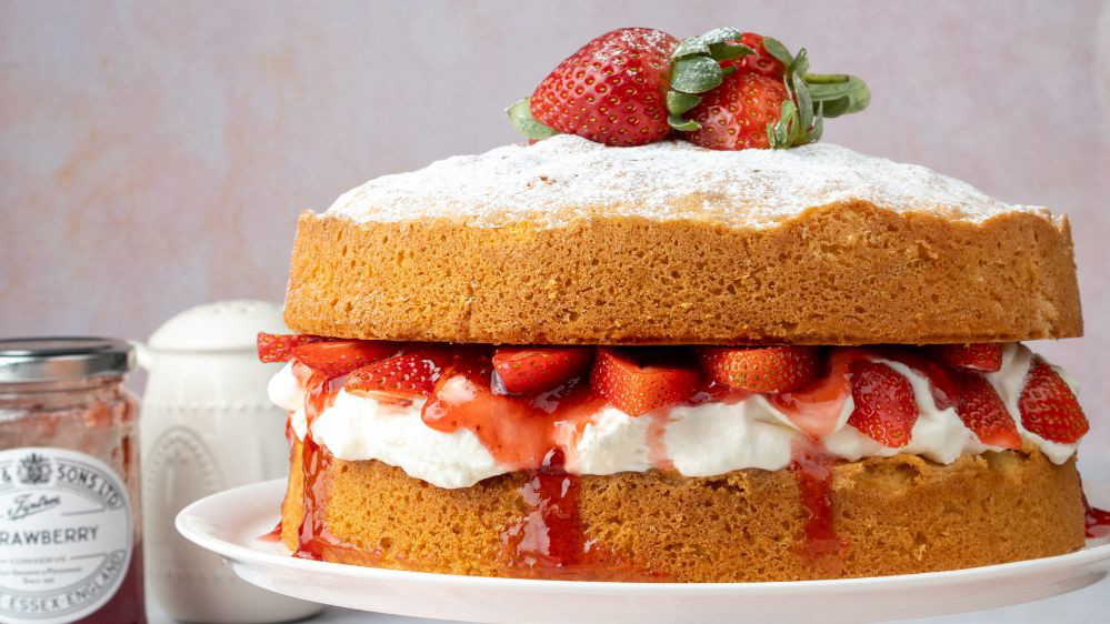 Image of a lavish Victoria Sponge filled with Strawberries and Cream next to a jar of Wilkin & Sons Tiptree Strawberry Jam