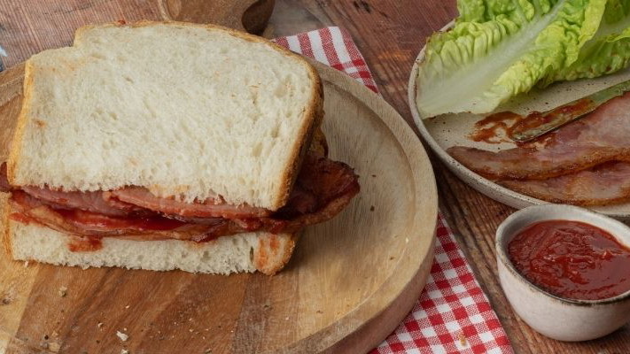 A bacon sandwich on white bread with a small pot of Tiptree Tomato Ketchup and lettuce off to one side.