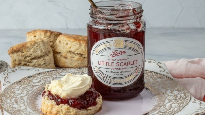 A scone with Jam and Clotted Cream in the correct order next to a jar of Tiptree Little Scarlet Strawberry Conserve