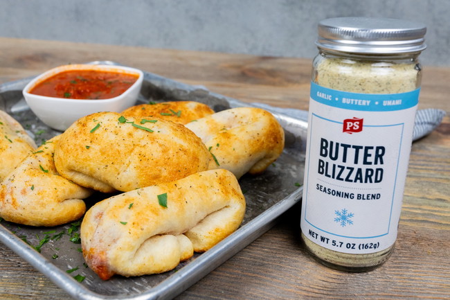 Butter Blizzard Pizza Rolls Golden brown baked pepperoni pizza rolls filled with melted mozzarella and pepperoni, brushed with Butter Blizzard seasoned butter. Displayed on parchment paper with a side of warm pizza sauce for dipping and a jar of PS Seasoning Butter Blizzard in the background.