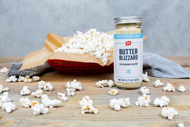 Butter Blizzard Popcorn Basket of freshly popped popcorn seasoned with PS Seasoning Butter Blizzard, with popcorn scattered on a wooden surface and a jar of Butter Blizzard seasoning in the foreground. Simple garlic butter snack made with stovetop popcorn.