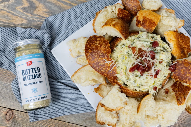 Butter Blizzard Bacon Spinach Artichoke Dip Warm spinach, bacon, and artichoke dip served in a crispy bread bowl, topped with shredded parmesan and surrounded by torn bread pieces. A jar of PS Seasoning Butter Blizzard is placed beside the dish on a striped napkin. Butter Blizzard spinach artichoke dip, bacon spinach dip recipe, PS Seasoning party dip, creamy artichoke bread bowl dip, garlic butter dip with bacon, baked spinach dip appetizer, savory cheese dip for bread