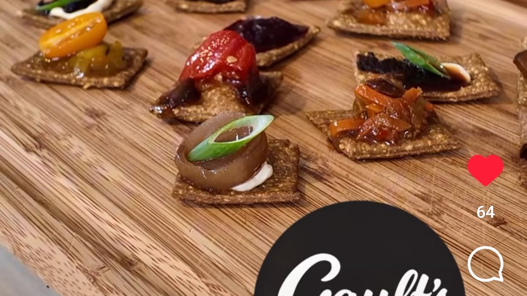 Square black garlic crackers with morsels of chutney and tomatoes on top arranged on a wooden board