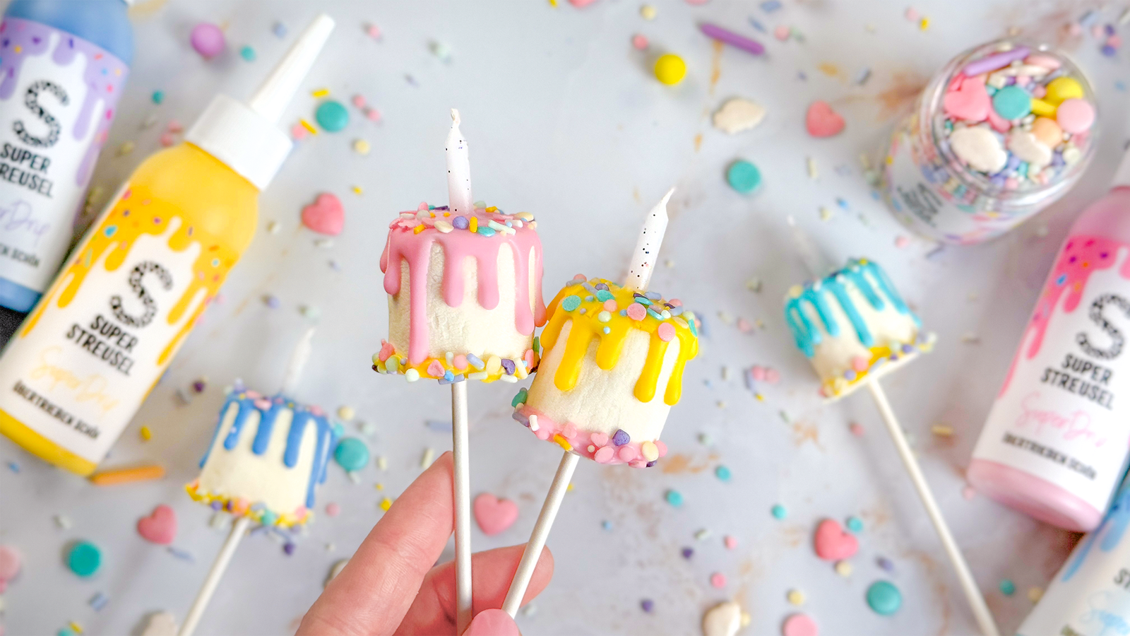 GeburtstagsTorten Pops aus Marshmallows am Cakepop-Stiel, dekoriert mit buntem Pastell-SuperDrip als Drip-Look, bestreut mit StreuselMix PastellKarussell und mit kleiner Geburtstagskerze in der Mitte.
