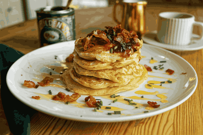  stack of fluffy pancakes topped with crispy bacon, chorizo, chili jam, and a drizzle of Lyle’s Golden Syrup, served on a white plate for a sweet-and-savory breakfast.