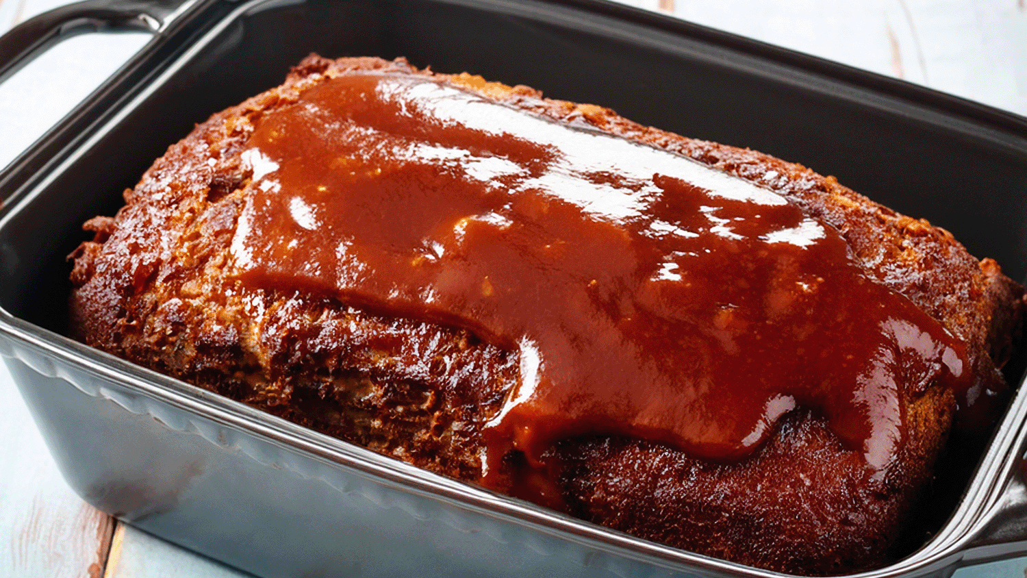 A close-up, high-angle shot of a freshly baked bison meatloaf in a dark rectangular loaf pan, resting on a light blue rustic wooden surface. The meatloaf is generously topped with a thick, glossy, reddish-brown BBQ sauce glaze that drips slightly down the sides.