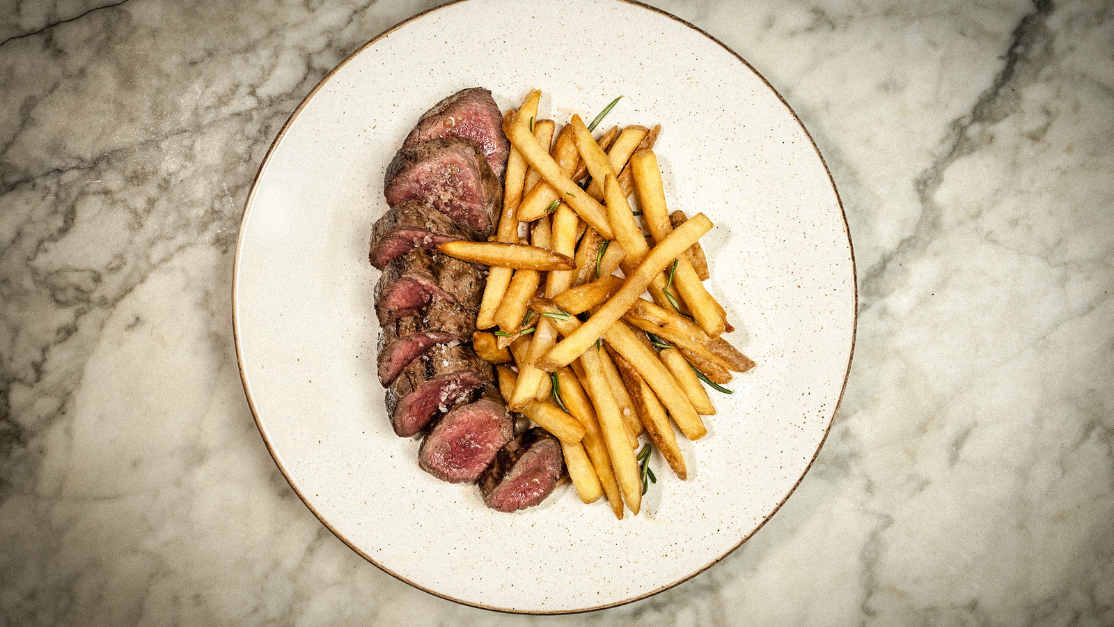 A plate with sliced venison striploin steak with chips on a marble countertop.
