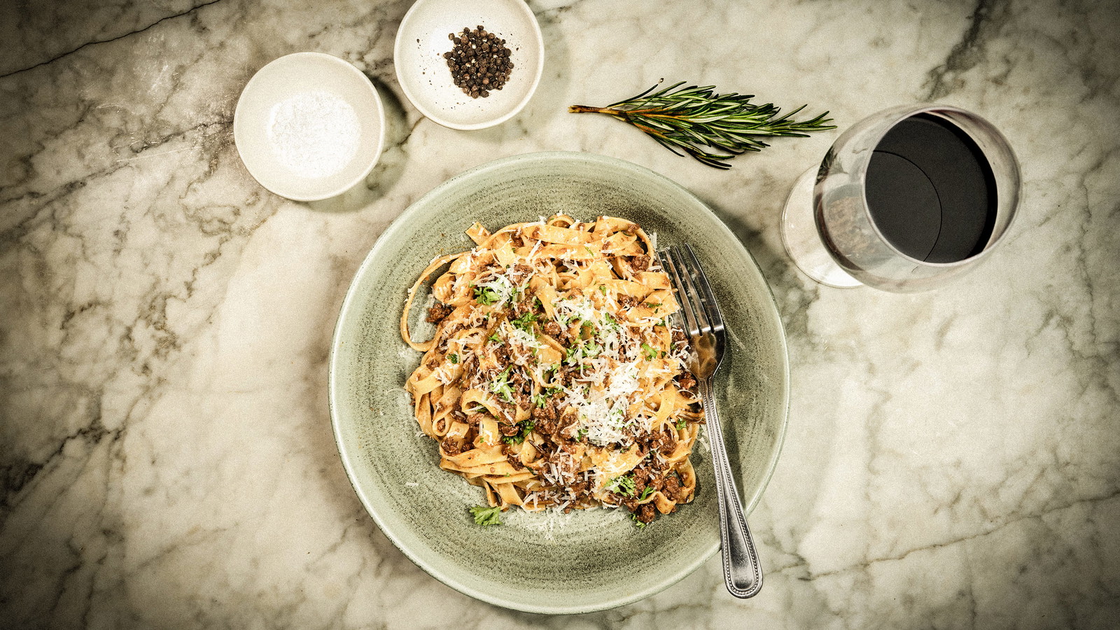 A bowl of pappardelle with venison ragu sprinkled with parmesan on a marble table with a glass of red wine.