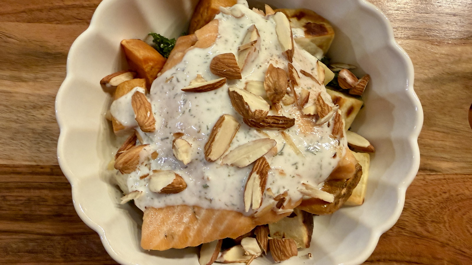 White scalloped bowl containing a salmon bowl with kale, roasted sweet potatoes, Dilliscious sauce and sliced almonds on top