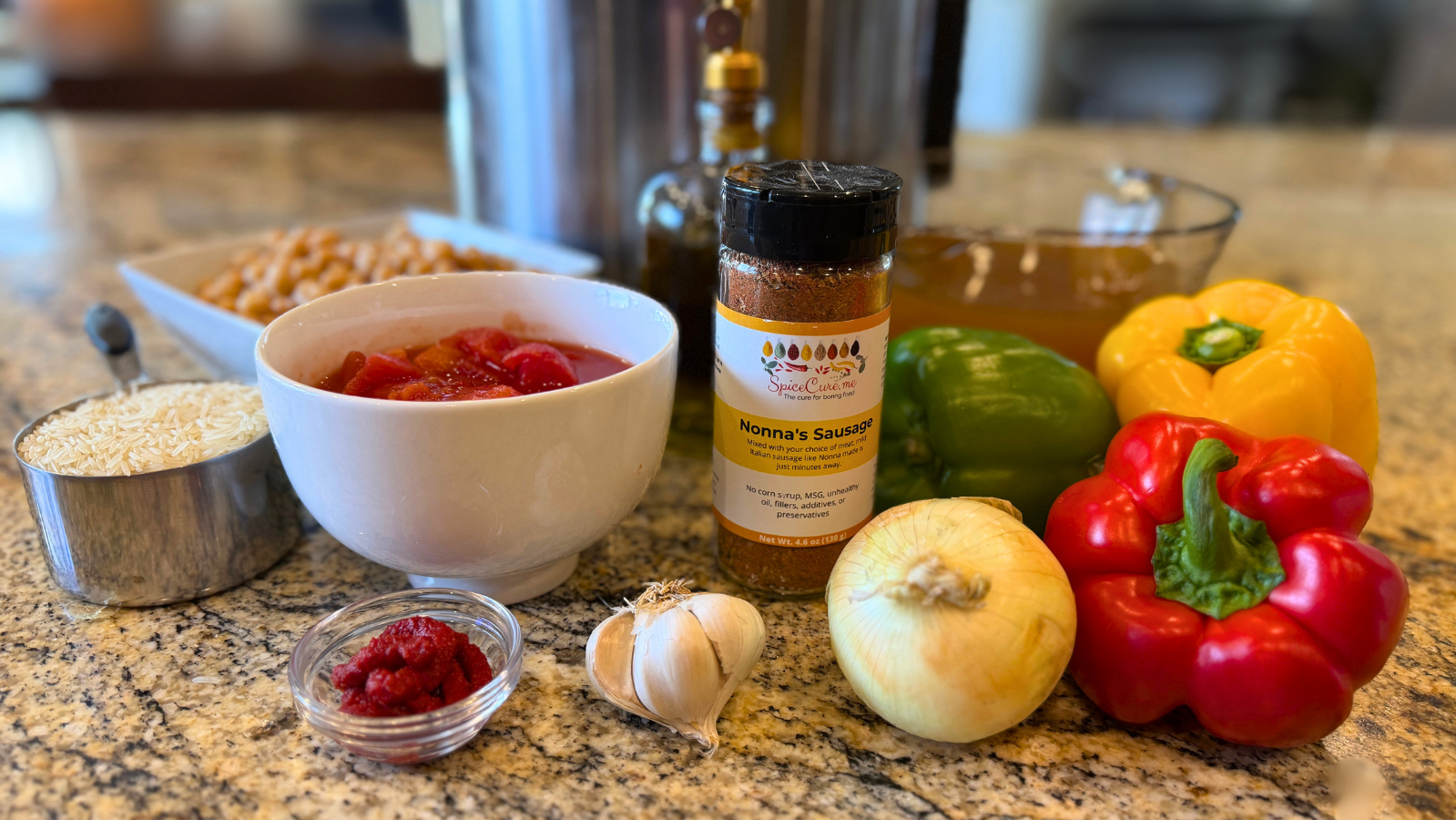 Colorful Instant Pot chickpeas and rice ingredients arranged on a granite countertop: basmati rice in a metal measuring cup, a white bowl of tomatoes, a small glass dish of tomato paste, garlic bulbs, a yellow onion, red, yellow, and green bell peppers, a glass measuring cup of vegetable broth, a rectangular bowl of chickpeas, a bottle of olive oil, and a jar of Nonna’s Sausage Italian seasoning in front of an Instant Pot