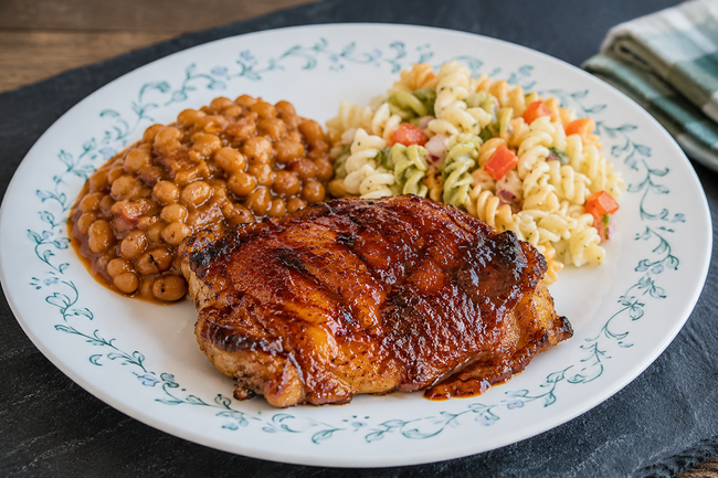 Rush to Table smoked barbecue chicken thigh finished with a caramelized barbecue glaze, served alongside baked beans and pasta salad on a classic Corelle-style dinner plate.