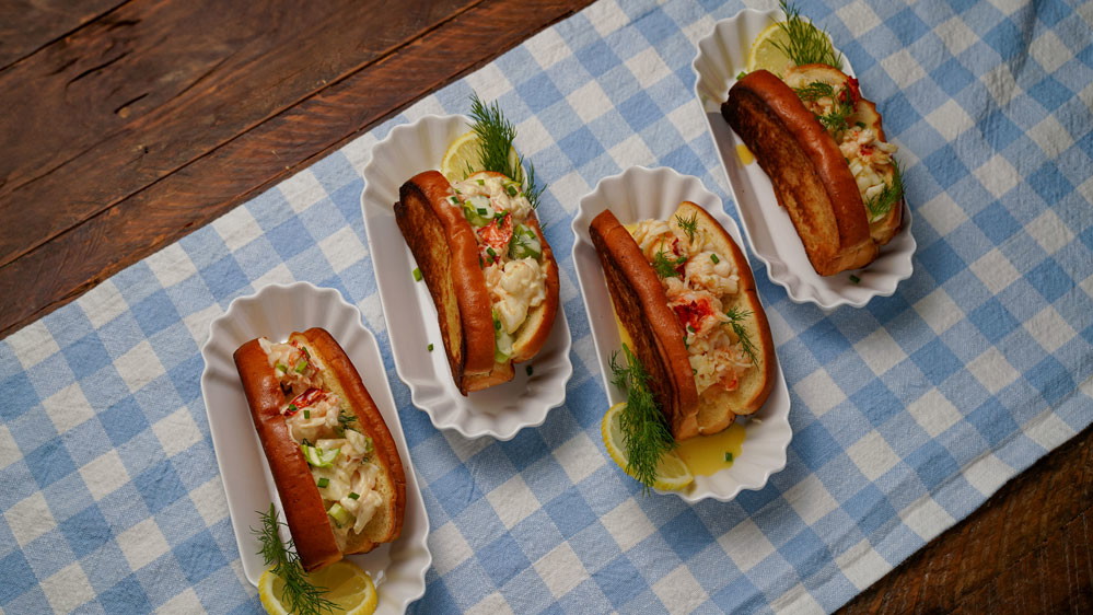 Four lobster rolls on a blue and white checkered table cloth