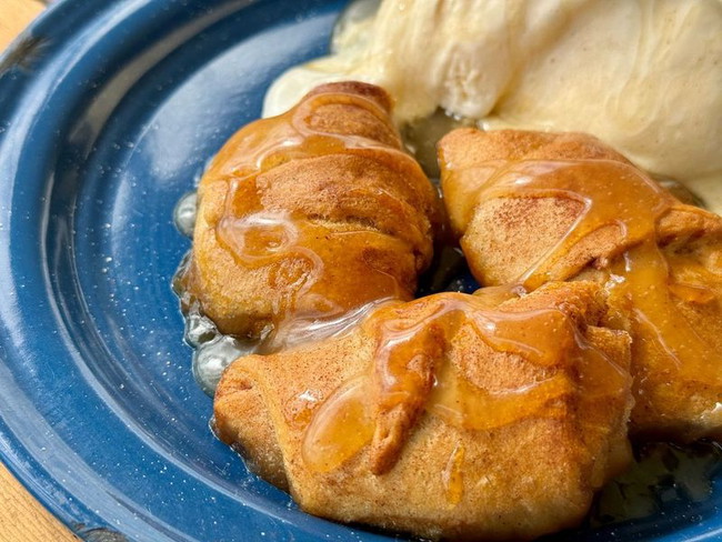 Image shows a blue plate with three apple dumplings, sprinkled with cinnamon and sugar and drizzled with sauce, next to vanilla ice cream.  
