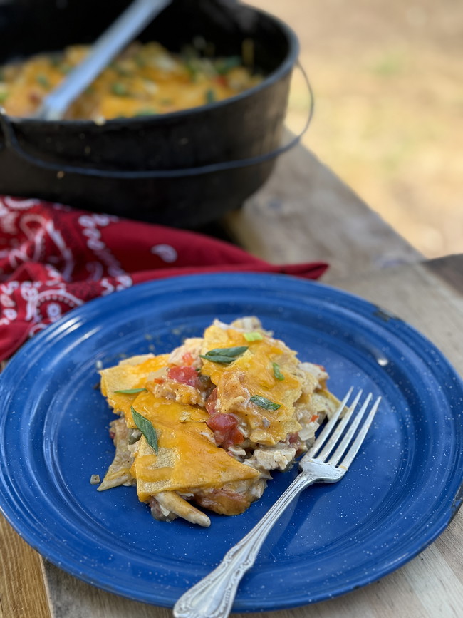A blue plate with a serving of casserole made with chicken, tortilla chips, cheese sitting on a table in front of a cast iron dish with the rest of the casserole.