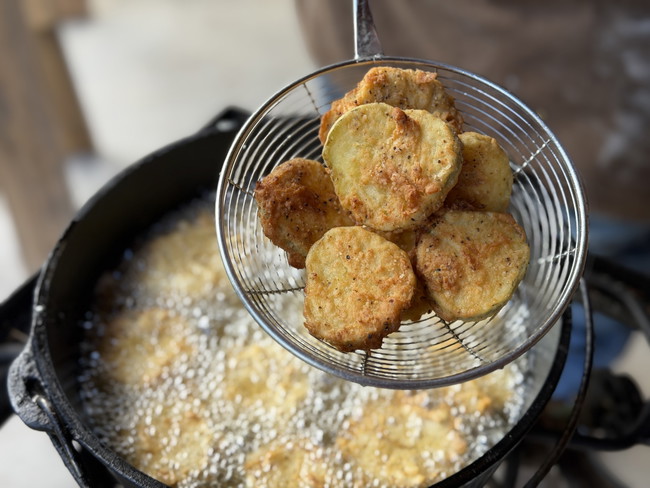 A basket of battered and fried pickle, held in a wire strainer over a cast iron pot of sizzling oil.
