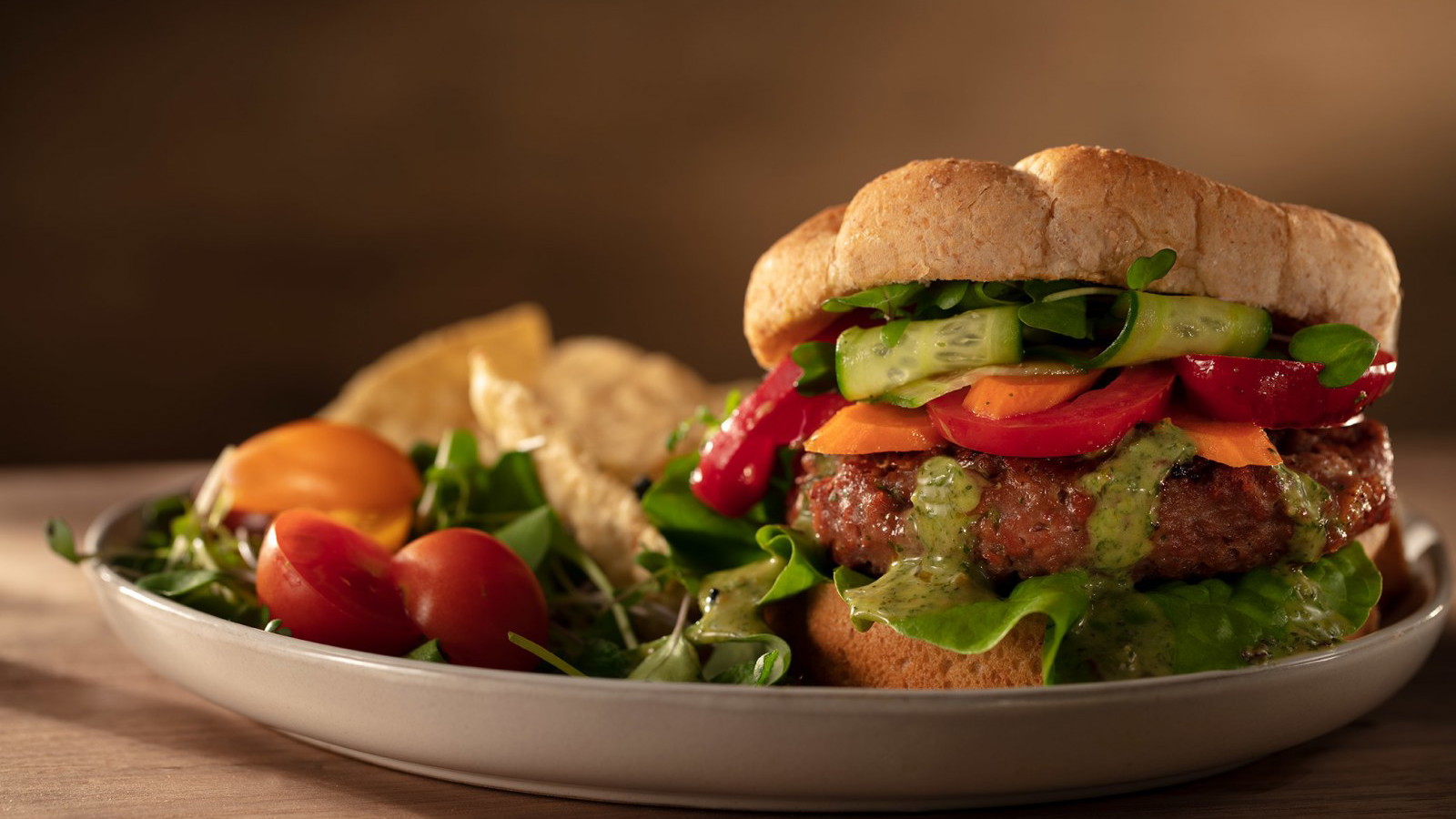 Veggie burger on a whole wheat bun with cilantro mint oo’mämē dressing, pickled vegetables, lettuce and tomato with a small salad and corn chips